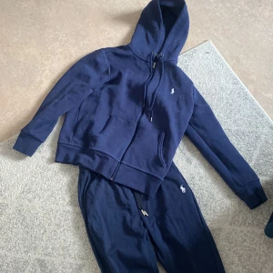 polo Ralph Lauren tracksuit - Snygg mörkblå tracksuit/dress från Polo Ralph Lauren med elastisk midja och dragsko. Byxorna har en liten broderad logga på sidan och muddar vid bensluten för en bekväm passform. Likadant med zip up koftan priset kan diskuteras använd 3 gånger som max