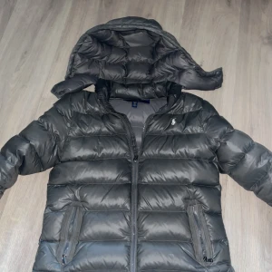 Grå dunjacka från Ralph Lauren - Säljer en snygg grå dunjacka från Ralph Lauren med huva och dragkedja. Jackan har långa ärmar och två praktiska fickor med dragkedjor. Perfekt för kyliga dagar.