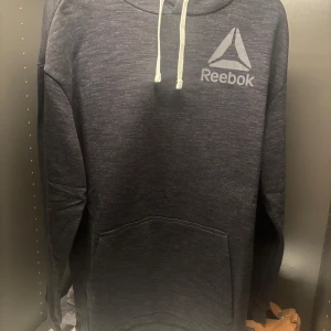 Mörkgrå hoodie från Reebok - Säljer en stilren mörkgrå hoodie från Reebok med logga på bröstet. Tröjan har en klassisk känguruficka och justerbar huva med snören. Perfekt för en avslappnad look.