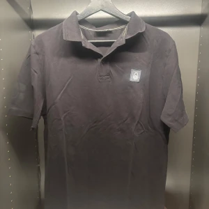 Svart pikétröja från Sail Racing - Snygg svart pikétröja från Sail Racing med korta ärmar och klassisk krage. Tröjan har en diskret logotyp på bröstet och är perfekt för en stilren look.