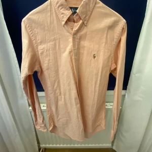 Rosa skjorta från Ralph Lauren - Snygg rosa skjorta från Ralph Lauren i storlek S. Skjortan har långa ärmar och en klassisk krage med knappar. Den är broderad med den ikoniska logotypen på bröstet. Perfekt för en stilren look.Tröjan är beige och rosa färg🔥🍾