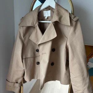 Beige trenchcoat - Snygg beige trenchcoat med dubbelknäppning och klassisk krage. Jackan är croppad och har långa ärmar med knappar vid ärmsluten..