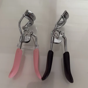 Ögonfransböjare med rosa och svarta handtag - Två ögonfransböjare med bekväma handtag i rosa och svart. Perfekta för att ge dina fransar en vacker böj och lyft. Metallisk finish med robust design för enkel användning.