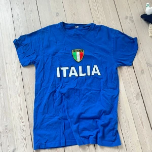 Blå t-shirt med Italienskt emblem - Säljer en blå t-shirt med texten 'ITALIA' och ett Italienskt emblem på bröstet. Perfekt för fotbollsfans eller Italien-älskare. T-shirten har en klassisk passform och korta ärmar.