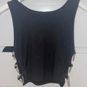 Svart ribbad topp med kedjedetaljer - Snygg svart ribbad topp med unika kedjedetaljer på sidorna. Toppen är ärmlös och har en tight passform, perfekt för en trendig look. Kedjorna ger en extra touch av stil och gör den till ett statement-plagg.