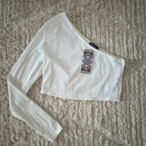 Vit oneshoulder topp - Snygg vit oneshoulder topp från Boohoo. Den är croppad och har en lång ärm på ena sidan. Perfekt för en trendig look. Oanvänd med etikett kvar.
