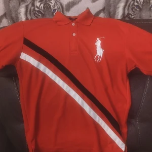 Röd pikétröja från Ralph Lauren - Snygg röd pikétröja från Ralph Lauren med klassisk krage och knappar. Tröjan har ett broderat vitt polo-spelarmärke på bröstet och en vit siffra '3' på ärmen. Diagonala ränder i svart och vitt ger en sportig touch.