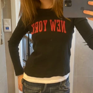 Svart långärmad tröja med röd text - Snygg svart långärmad tröja med röd text 'NEW YORK' på bröstet. Perfekt för en casual look med jeans. Tröjan har en normal passform och är gjord i ett bekvämt material.