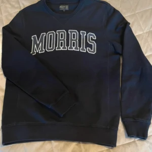 Mörkblå sweatshirt från Morris - Säljer en stilren mörkblå sweatshirt från Morris med deras logga i vitt på bröstet. Tröjan har långa ärmar och en klassisk rund halsringning. Perfekt för en avslappnad och trendig look.