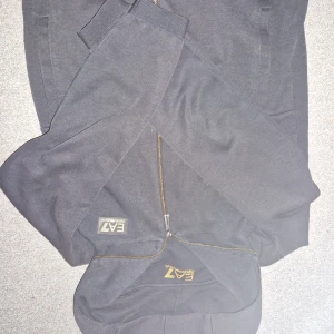 Svart hoodie från Emporio Armani - Säljer en stilren svart hoodie från Emporio Armani med dragkedja framtill. Den har en klassisk design med EA7-logga på bröstet och långa ärmar. Perfekt för en avslappnad look.