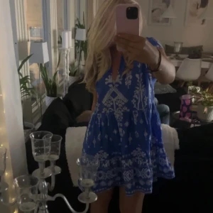 Zara klänning - Charmig blå kortklänning med ett vackert vitt mönster. Klänningen har en avslappnad passform och en v-ringad hals. Perfekt för sommarens varma dagar.