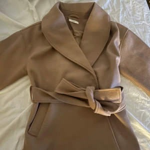 Kort kappa - Beige/brun kappa från GinaTricot. Storlek S. Kort modell med knytskärp i midjan. Assnygg nu till våren 🌸 Aldrig använd. Pris kan diskuteras!!