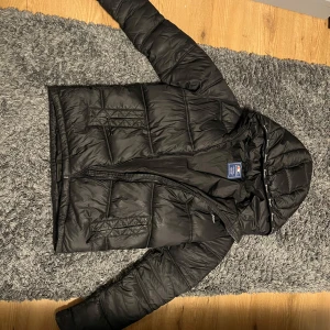 Svart pufferjacka från Champion - Säljer en svart pufferjacka från Champion med broderad logga på ryggen. Jackan har en dragkedja framtill och är långärmad med en skön huva. Perfekt för kyliga dagar.