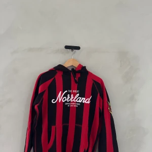 Svart och röd randig hoodie från The Great Norrland - Säljer en cool svart och röd randig hoodie från The Great Norrland. Den har en stor luva och en bekväm passform med känguruficka framtill. Perfekt för höst och vinter när du vill hålla dig varm och stilren. Texten 'Norrland' är tryckt på bröstet. Passar både till vardags och chillkvällar.