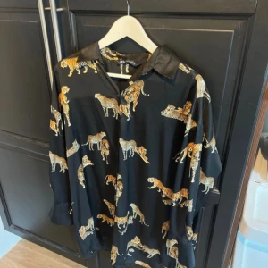 Svart skjorta med tigertryck från Zara - Säljer en cool svart skjorta från Zara i storlek XS. Den har ett unikt tigertryck i gult och orange som verkligen sticker ut. Skjortan har långa ärmar och en klassisk krage. Perfekt för att ge din outfit en djärv touch! 🐯