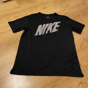 Svart t-shirt från Nike - Säljer en svart Nike t-shirt i Dri-FIT material, perfekt för träning. Den har en stor vit Nike-logga på framsidan och är kortärmad. Passar ungdomar i åldern 13-15 år. Skön och andas bra, idealisk för sportiga aktiviteter.