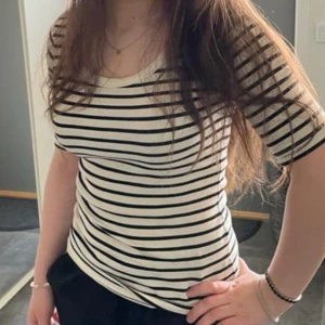 Randig topp i svart och vit - Säljer en snygg randig topp i svart och vit. Den har en tight passform och korta ärmar, perfekt för både vardag och fest. Materialet känns mjukt och bekvämt mot huden. Passar bra till jeans eller kjol för en stilren look.