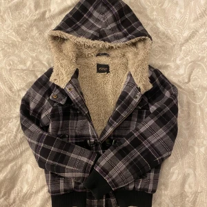 Svart och grå rutig jacka med teddyfoder - Säljer en supermysig svart och grå rutig jacka från Eve. Den har ett varmt teddyfoder och en huva som gör den perfekt för kyliga dagar. Jackan har både knäppning med snygga knappar och dragkedja. Perfekt för höst och vinter!
