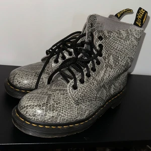 Grå mönstrade boots från Dr. Martens - Säljer ett par grå mönstrade Dr. Martens boots i bra skick. De har en cool ormskinnslook och klassisk gul söm. Perfekta för höst och vinter med slitstark sula och snörning. Passar både till vardags och fest!