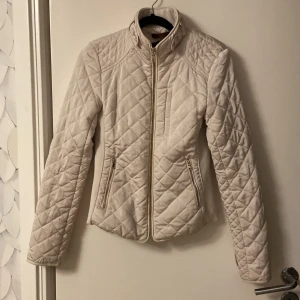 Beige quiltad jacka med dragkedja från Zara - Säljer en snygg beige quiltad jacka i nyskick. Den har en dragkedja framtill och två fickor med dragkedjor. Jackan har långa ärmar och en hög krage med knappar och fina detaljer. Perfekt för höst och vår!