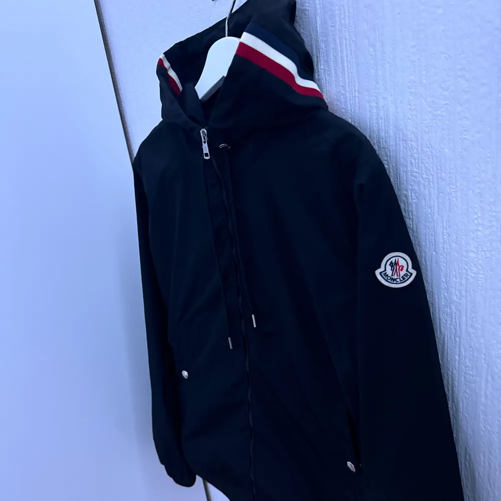 Säljer en snygg svart jacka från Moncler. Den har en dragkedja framtill och en huva med röd och vit randdetalj. Perfekt för höst och vår! Jackan har långa ärmar och Moncler-loggan på ärmen. Passar både till vardags och lite finare tillfällen.. Jackor.