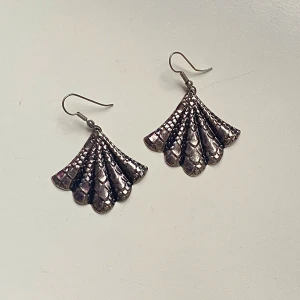 Silvriga örhängen i fläktform - Säljer ett par unika silvriga örhängen i form av en fläkt. De har en vacker textur som påminner om fjäll och ger en elegant look. Perfekta för att piffa upp vilken outfit som helst, oavsett om det är till vardags eller fest. Örhängena är i nyskick och redo att bäras av någon som älskar att sticka ut!