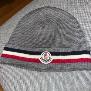 Moncler mössa - Säljer en grå Moncler mössa i storlek one size. Den är i fint skick och har den klassiska Moncler-loggan framtill samt Frankrikes flagga. Perfekt nu inför vintern. Mössan är bekväm och varm, tillverkad av högkvalitativa material.