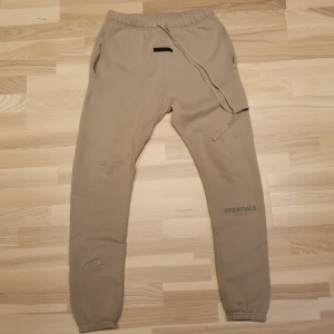 Essentials mjukis byxor stl S - Essentials fear of god Mjukisbyxor självklart äkta Stolek S  Beige bomullsblandning fleece konsistens resår i midjan med dragsko gummerad logotyp logotyptryck vid benet två sidofickor med slitsar elastiska anklar