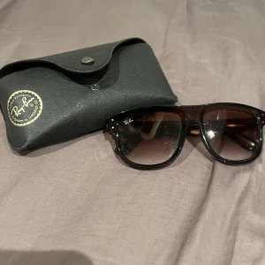 Solglasögon från Ray-Ban - Säljer ett par snygga Ray-Ban solglasögon med brun toning och klassisk design. De kommer med ett svart fodral som har Ray-Ban-loggan i guld. Perfekta för både sommar och vår, dessa glasögon ger en stilren look och skyddar dina ögon mot solen. Passa på att köpa dem nu!