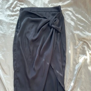 Svart omlottkjol med knytband - Snygg svart omlottkjol i satin med knytband framtill. Kjolen har en elegant slits och en diskret dragkedja på sidan. Perfekt för både fest och vardag! 🖤