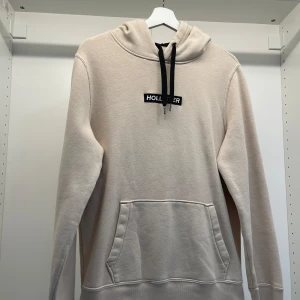 Beige hoodie från Hollister - Säljer en snygg beige hoodie från Hollister i storlek M. Den har en klassisk design med en känguruficka och svarta dragsnören vid huvan. Perfekt för en avslappnad stil under höst och vår. Supermjuk och bekväm!