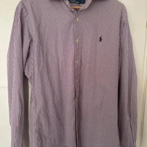 Randig skjorta från Ralph Lauren - Säljer en klassisk långärmad skjorta från Ralph Lauren i storlek L. Skjortan är vit och lila randig med knappar framtill och det ikoniska broderade logotypen på bröstet. Tillverkad i 100% bomull, perfekt för både vardag och fest. 🕺