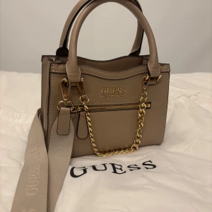 Beige handväska från Guess - Säljer nu min skitsnygg handväska i beige färg från Guess. Jag har använt den kanske 3-4ggr så den är i nyckick. Den har guldfärgade detaljer och en avtagbar axelrem med Guess-logga. Väskan har flera fack och en dragkedja framtill. Perfekt för både vardag