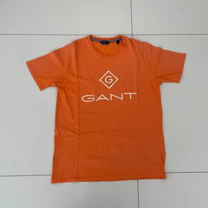 Orange t-shirt från GANT - Säljer en snygg orange t-shirt från GANT i bomull. Den har en klassisk passform med korta ärmar och en rund halsringning. Perfekt för sommardagar eller avslappnade tillfällen. GANT-loggan är tryckt i vitt på framsidan. Skicka ett meddelande vid intresse! 🧡