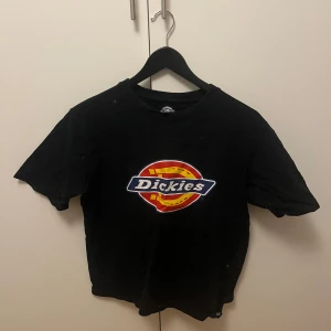 Svart t-shirt från Dickies - Säljer en svart t-shirt från Dickies med deras ikoniska logga på bröstet.  T-shirten är kortärmad och i bomull, vilket gör den skön att bära till vardags. Katt finns i hemmet men den kommer vara rollad och tvättad innan den skickas 
