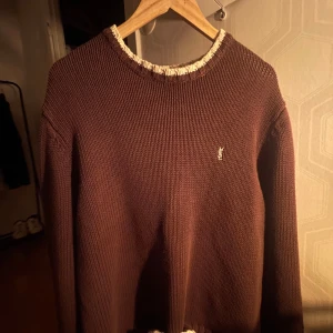Brun stickad tröja från Yves Saint Laurent - Säljer en snygg brun stickad tröja från Yves Saint Laurent. Den har en krämfärgad ribbad kant vid halsen och ett litet broderat emblem på bröstet. Perfekt för höst och vinter, både stilren och bekväm. Passar till både vardag och fest! Nypris ca: 13 000kr