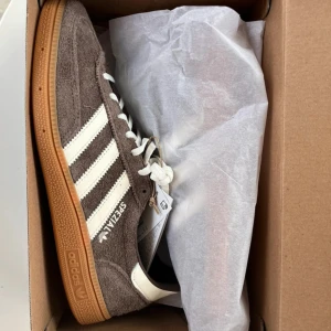 Adidas Spezial Handball Earth Strata - Säljer ett par snygga bruna Adidas sneakers i modellen Spezial Handball. De är i mocka med vita detaljer och har en klassisk gummisula. Colorway Earth Strata. Skorna är i nyskick och kommer i originalkartong. Storlek passar perfekt för en casual look.