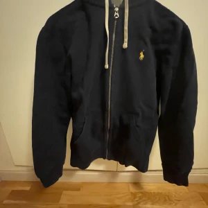 Mörkblå hoodie från Ralph Lauren - Säljer en snygg mörkblå hoodie från Ralph Lauren med dragkedja och vit dragsko. Den har en broderad logga på bröstet och är perfekt för en casual look. Passar bra till både jeans och joggers.