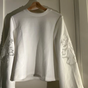 Vit tröja med spetsdetaljer - Säljer en stilren vit tröja med långa ärmar och vackra spetsdetaljer på ärmarna. Perfekt för en elegant och avslappnad look. Tröjan har en normal passform och är gjord i ett mjukt material.