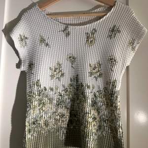 Säljer en vit topp med blommigt mönster i gröna och gula nyanser. Toppen har korta ärmar och en ribbad struktur som ger en unik look. Perfekt för en somrig stil!