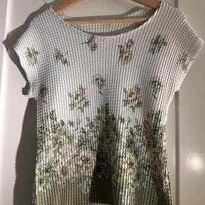 Vit blommig topp - Säljer en vit topp med blommigt mönster i gröna och gula nyanser. Toppen har korta ärmar och en ribbad struktur som ger en unik look. Perfekt för en somrig stil!