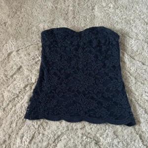 Mörkblå spets tubtopp - Snygg mörkblå tubtopp i spets med blommigt mönster. Perfekt för en stilren och elegant look. Toppen är axelbandslös och har en fin passform som framhäver figuren.