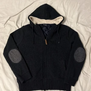 Stickad cardigan  - Säljer en svart stickad cardigan från Peacocks med dragkedja och mysigt fleecefoder i luvan. Den har ribbade muddar och snygga grå armbågslappar. Perfekt för kyliga dagar! 🖤