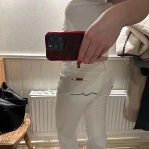 Vita jeans med detalj - Snygga vita jeans med hög midja och en detalj på bakfickan. De har en klassisk femficksdesign och stängs med dragkedja och knapp. Perfekta för en stilren look.