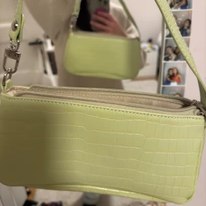 Ljusgrön axelväska i syntet - Snygg ljusgrön axelväska med krokodilpräglat mönster. Väskan har en dragkedja upptill och en justerbar axelrem. Perfekt för att ge en färgklick till din outfit!