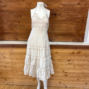 Vit klänning med pärldetaljer - Charmig beige klänning med halterneck och v-ringning, dekorerad med små pärlor i rött och guld. Kjolen har flera volanglager som ger en lekfull känsla. Perfekt för sommarens festligheter!