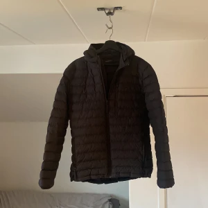 Svart quiltad jacka från Peak Performance - Snygg svart quiltad jacka från Peak Performance med huva och dragkedja. Perfekt för kyliga dagar med sin stilrena design och praktiska fickor. Jackan har långa ärmar och är tillverkad i ett lätt och bekvämt material.