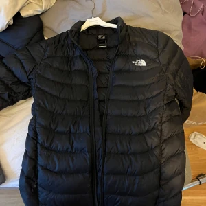 Svart dunjacka från The North Face - Säljer en svart dunjacka från The North Face. Jackan har en stilren design med dragkedja och långärmade ärmar. Perfekt för kyligare dagar och håller dig varm och bekväm. Passar både till vardags och äventyr. 🖤