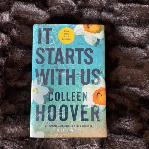 It Starts With Us - Boken 'It Starts With Us' av Colleen Hoover har en omslagsdesign med blå bakgrund och vita blommor med orangea detaljer. Den är en uppföljare till 'It Ends With Us' och är en populär roman av en bästsäljande författare.