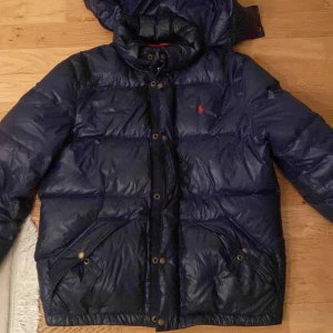 Blå dunjacka från Ralph Lauren - Säljer en snygg blå dunjacka från Ralph Lauren. Jackan har en quiltad design med en huva och knappar framtill. Den är perfekt för kalla dagar och har en röd insida som ger en cool kontrast. Märkesloggan syns på bröstet. Passar perfekt för vintern! 💙